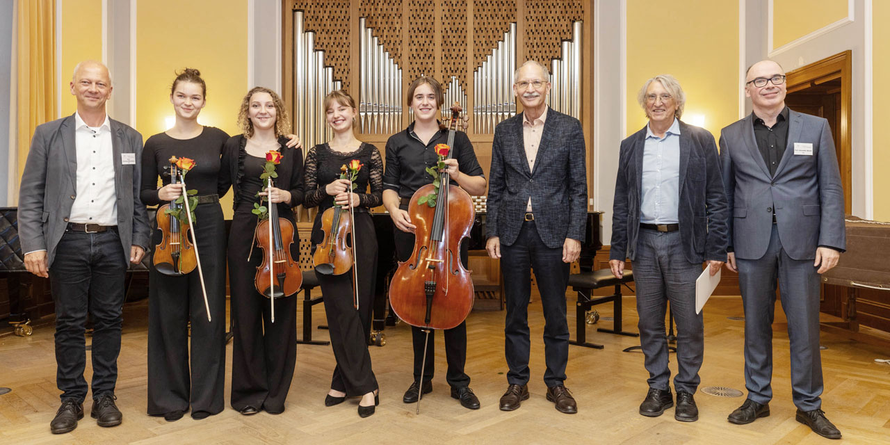 Vier ältere Männer in Anzügen stehen neben vier jungen Orchestermitgliedern, welche ihr Instrument in der Hand halten und ebenfalls ordentliche schwarze Kleidung tragen. Drei Jugendliche halten eine Geige und einer hält ein Cello.