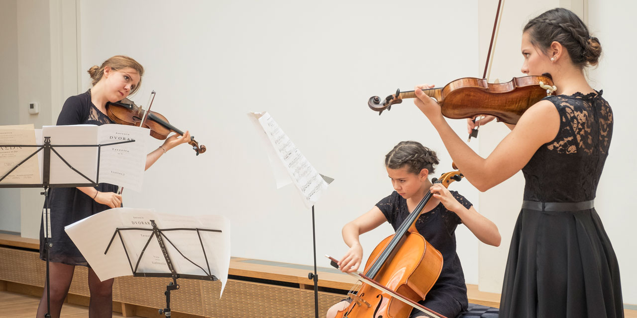 Drei Jugendliche tragen dunkelblaue Kleider. Zwei von ihnen spielen Geige und eine spielt Cello.