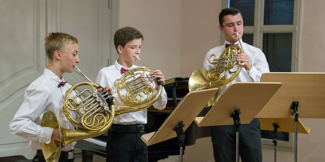 Drei Jugendliche tragen ein weißes Hemd und spielen Horn.