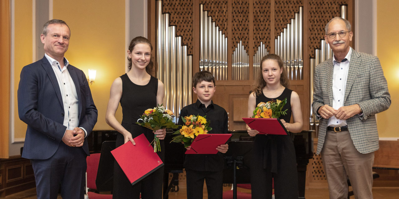 Zwei ältere Männer stehen neben drei Kindern, welche eine rote Mappe und einen Blumenstrauß in der Hand halten. Im Hintergrund ist eine Orgel zu sehen.