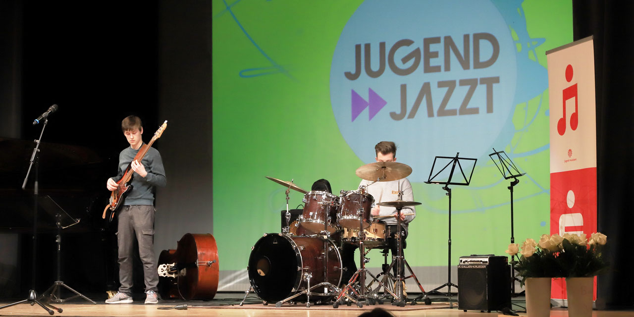 Ein Schlagzeuger und ein Bassist spielen gemeinsam auf einer Bühne. Im Hintergrund ist das Logo von JUGEND JAZZT an die Wand projiziert.