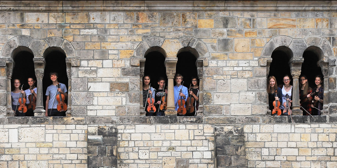 Jugendliche halten ihre Geigen in der Hand und schauen aus drei Fenstern hinaus. Die Fenster sind in einer alten großen steinwand eines Gebäudes und es gibt kein Glas. In zwei Fenstern stehen vier Jugendliche und in einem Fenster ganz links drei.