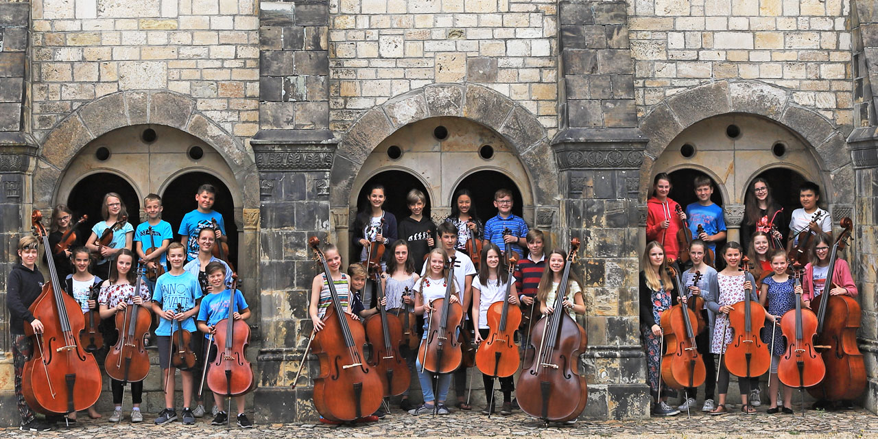Junge Cello und Kontrabass Spieler/-innen stehen im Vordergrund und dahinter junge Geigen und Bratschen Spieler/-innen in steinernen Torbögen.
