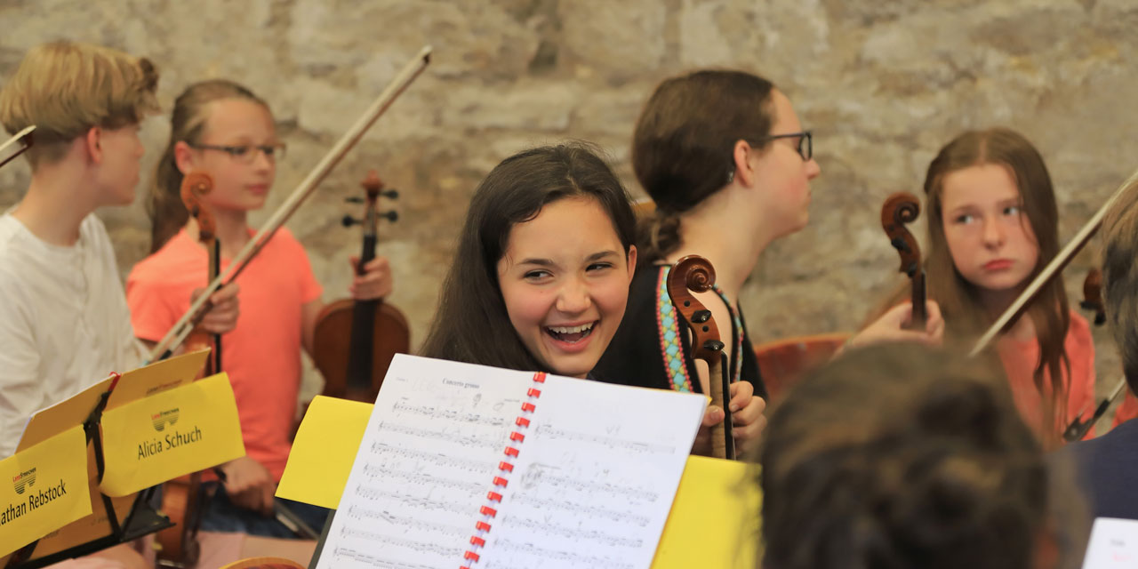 Ein Mädchen lacht jemanden an beim Proben, während im Hintergrund vier weitere Kinder miteinander reden. Eine Nahaufnahme von einem jungen Mädchen mit ihrem Cello.