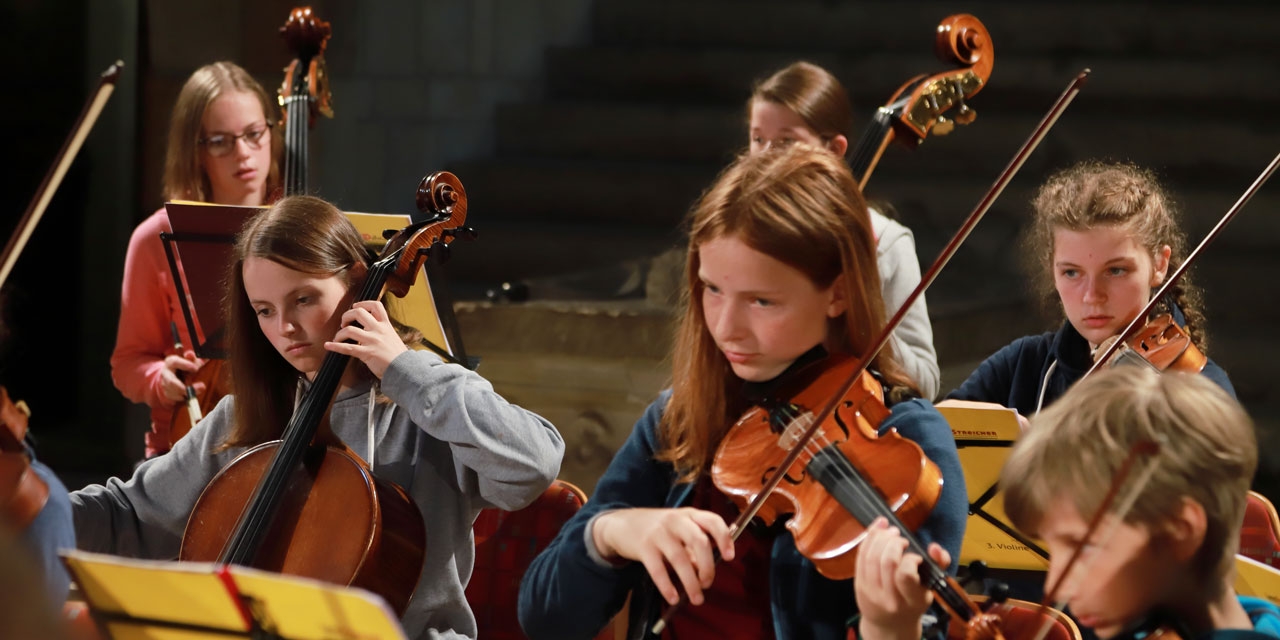 Mehrere Teilnehmer-/innen sind beim Spielen ihrer Instrumente zu sehen. Im Bildmittelpunkt befindet sich eine junge Bratscherin mit orangeroten Haaren und blauer Jacke, die konzentriert in die Noten schaut, neben ihr sitzt eine Cellistin in grauem Oberteil