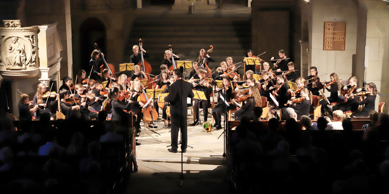 Die Landstreicher spielen 2018 in einer Kirche. Alle Kinder tragen schwarze Kleidung, die Notenmappen des jungen Streichorchesters sind auffällig gelb. Zentral im Bild steht der ebenfalls schwarz gekleidete Dirigent, davor sieht man die mit Zuschauern besetzten Kirchenbänke.