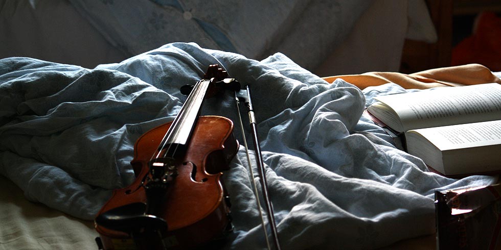 Eine Violine und ein offenes Buch liegen auf einer hellblauen Decke auf einem Bett.