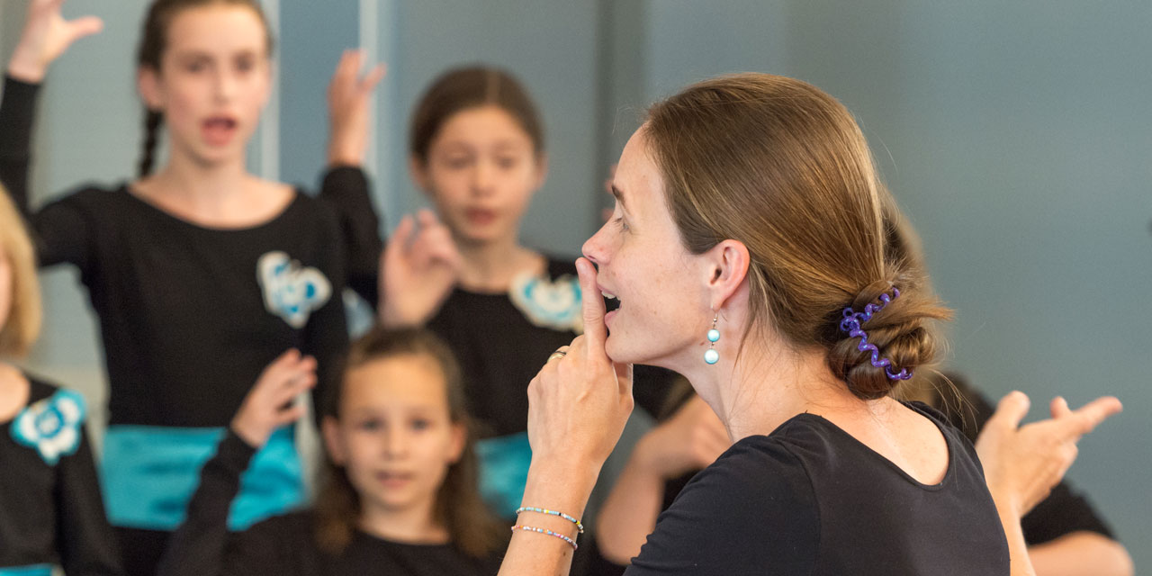 Eine Chorleiterin legt den Finger auf die Lippen, im Hintergrund ihr Chor