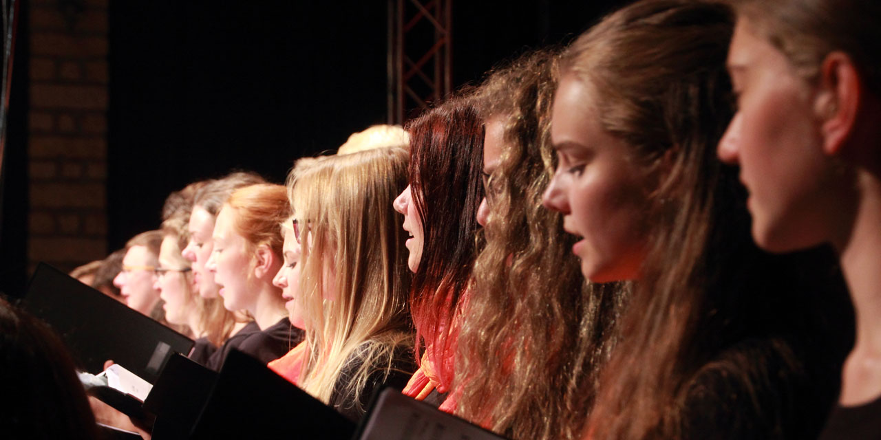 Im Vordergrund sieht man mehrere in einer Reihe stehende ältere Teenagerinnen, die in einem Chor singen und eine Notenmappe halten. Weiter hinten setzt sich die Reihe auch mit Jungen fot, vor und hinter ihnen stehen ebenfalls junge Mitglieder des Chors.