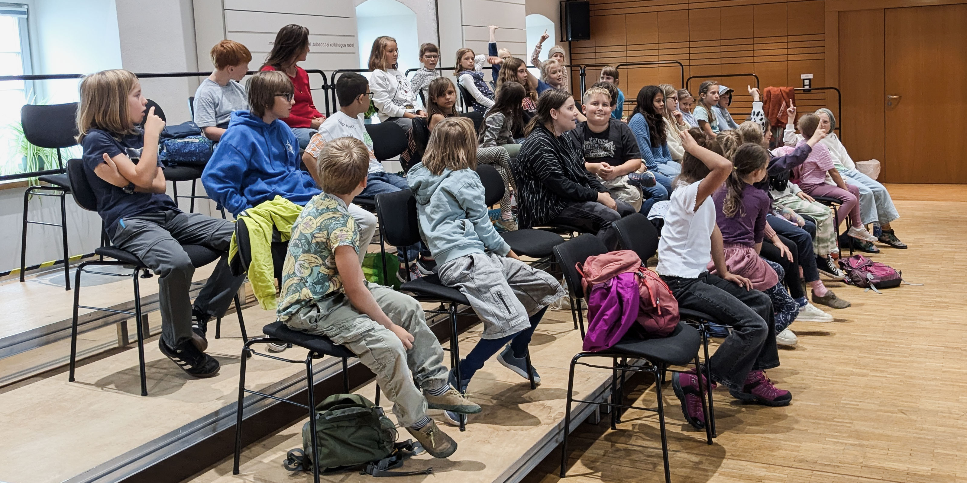 Zahlreiche Kinder sitzen auf vier Podeststufen in der Landesmusikakademie Sachsen. Einige Kinder melden sich – eine Unterrichts- oder Probensituation